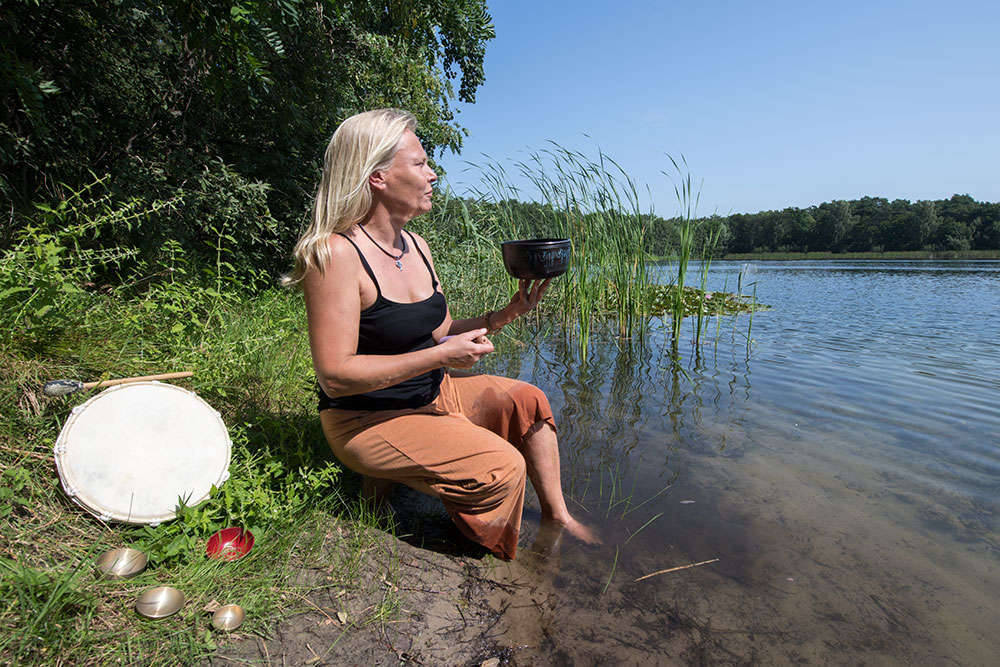 Person mit Klangschale und Trommel sitzend am See
